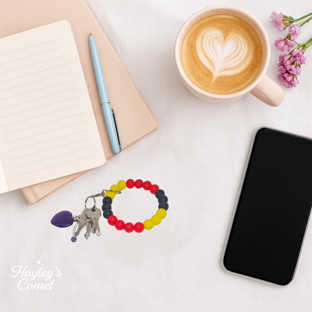 Colorful beaded keychain with keys on a desk beside a cup of coffee, smartphone, and notebook.